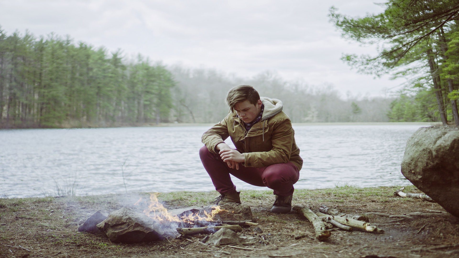A man in an olive green jacket is crouching down next to the edge of a lake as he examines his camp fire.