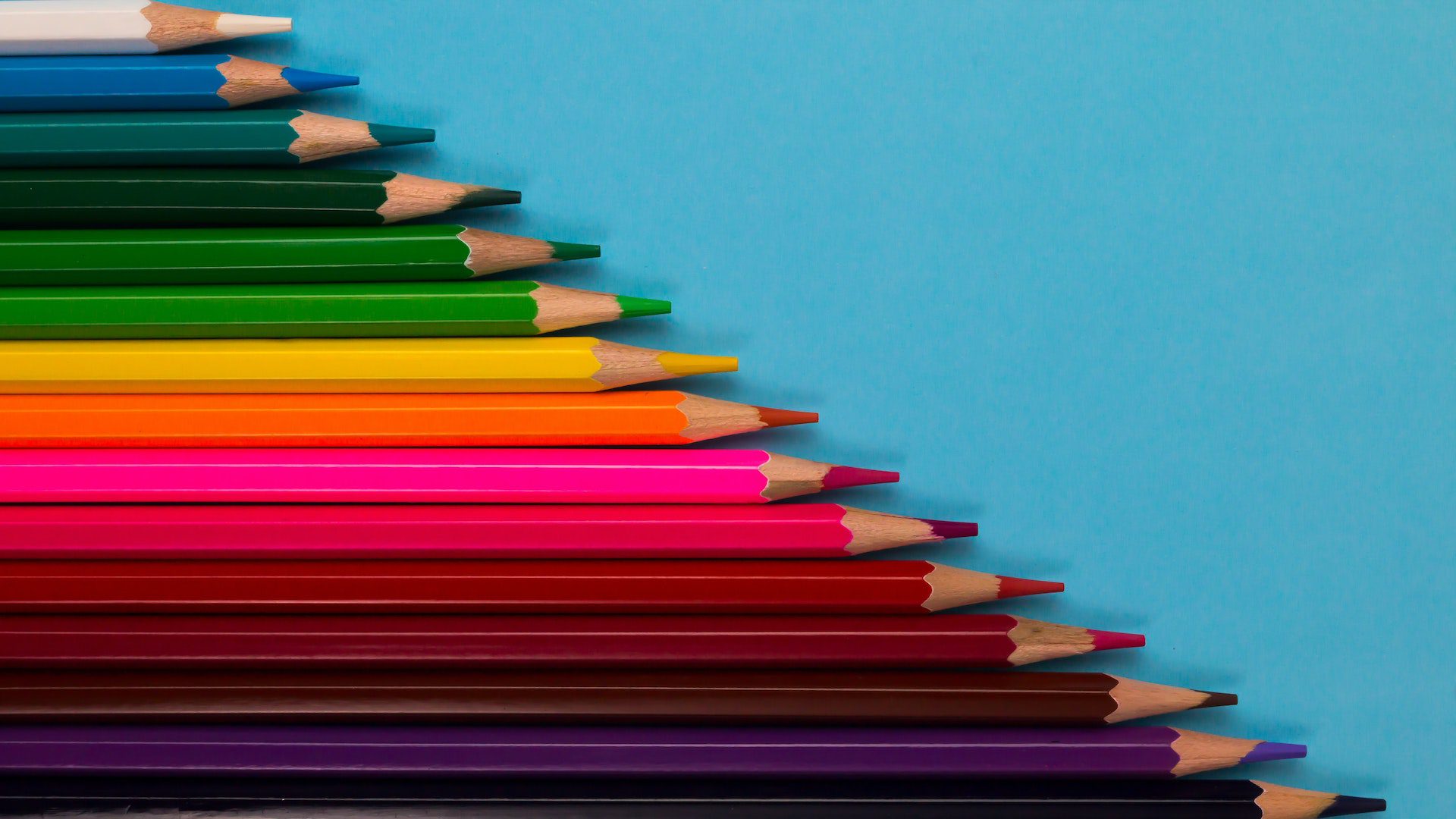 A variety of color pencils arranged in a stair-step pattern on a blue piece of paper.