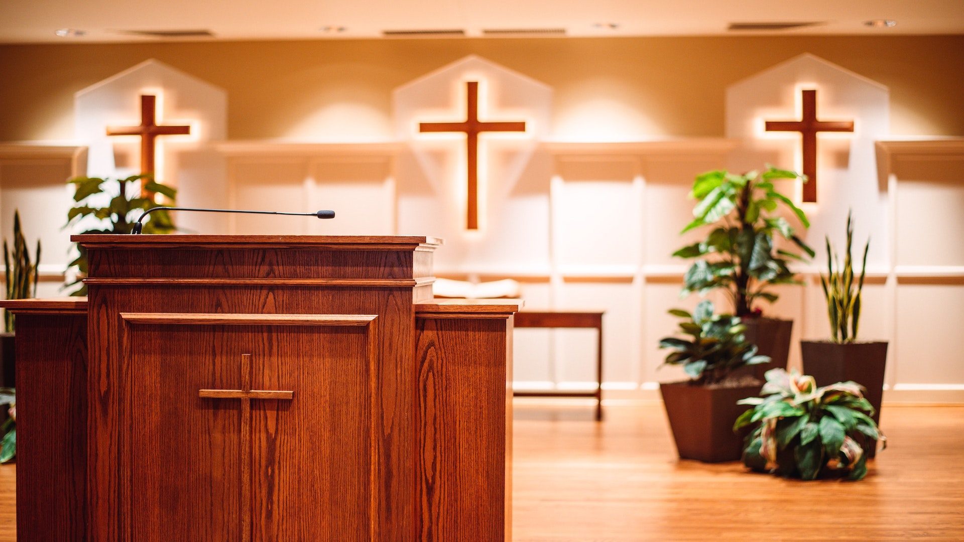 A brown wooden pulpit with a cross. There are also a variety of potted plants around the pulpit.