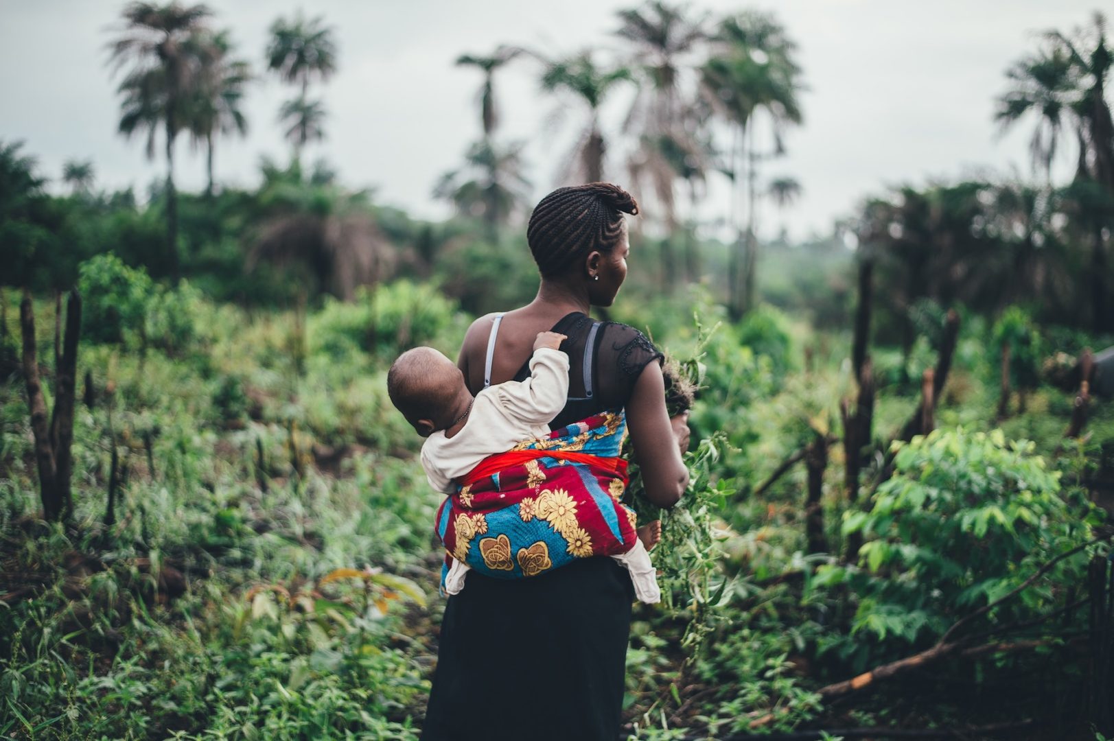 An African woman walks through the jungle with her child on her back.