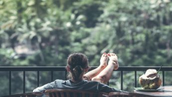 Back of a woman sitting outside with her feet resting on a fence.