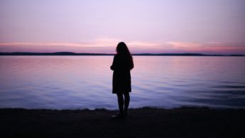 Silhouette of a woman standing in front of a lake during a pink and purple sunset.