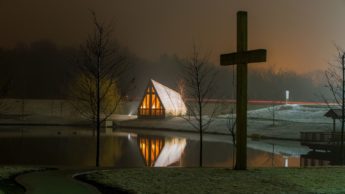 Cross on grass in front of a lake and church at night.