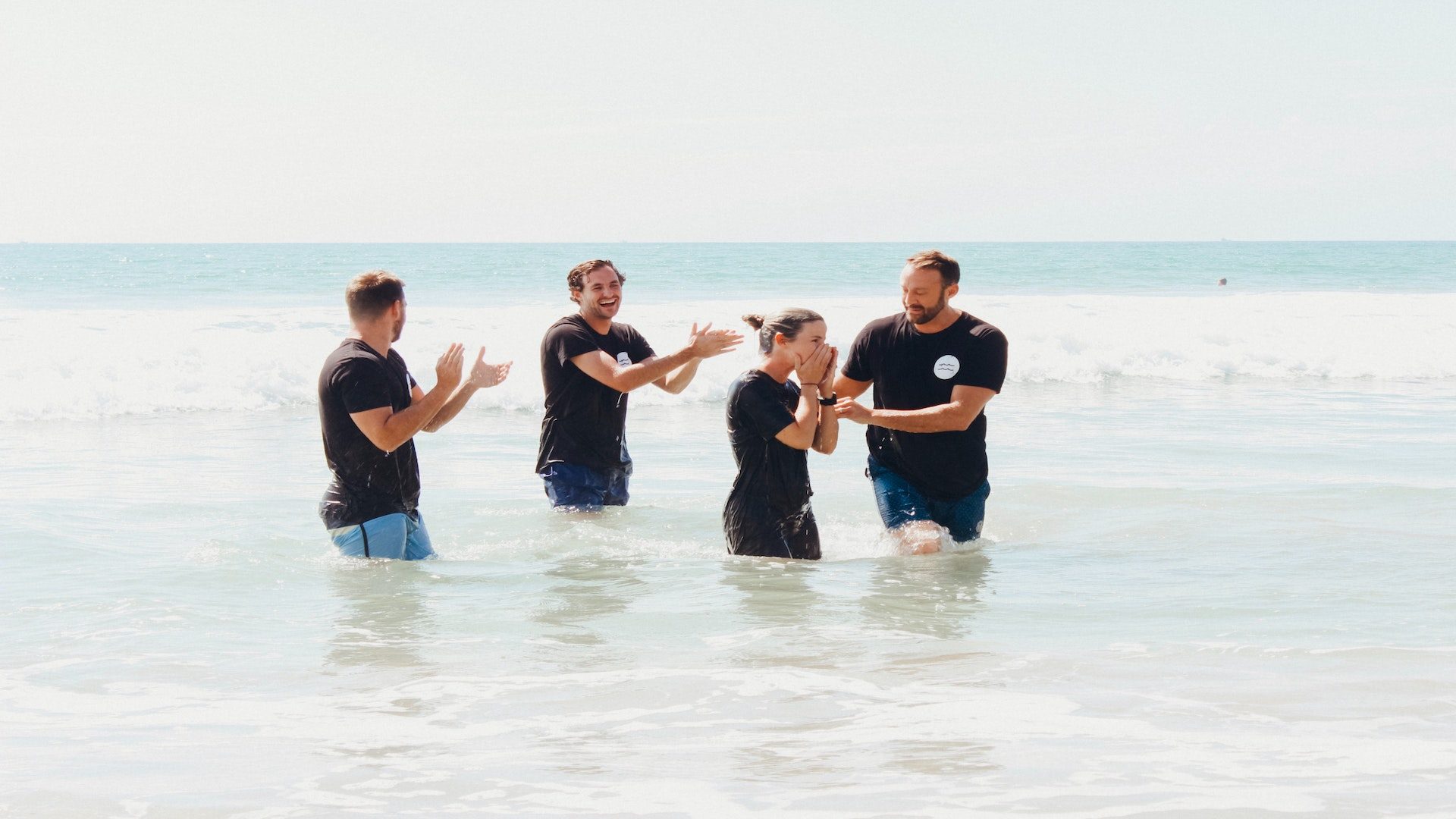 Four people in black t-shirt standing in an ocean clapping.
