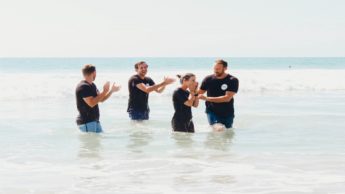 Four people in black t-shirt standing in an ocean clapping.