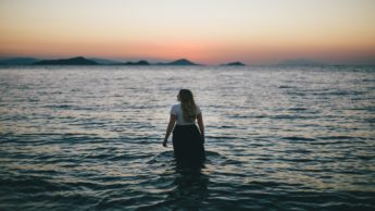 Woman in a white shirt and black skirt standing in water during a sunset.