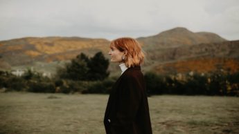 Woman in a black coat walking in a green field near mountains.