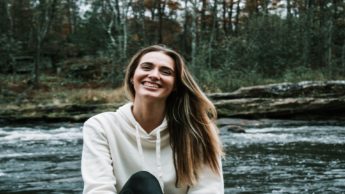 Woman inWoman in a white sweatshirt sitting in front of a stream. a white shirt standing in front of trees.