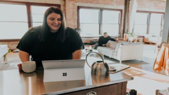 Woman standing at a table with a mug and laptop.