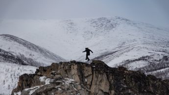Person jumping on brown, rocky mountain next to snowy mountains.