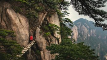 Man wearing a red jacket climbing up stairs on the side of a cliff.