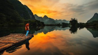 Man standing on a bamboo raft in water next to mountains during a sunset.