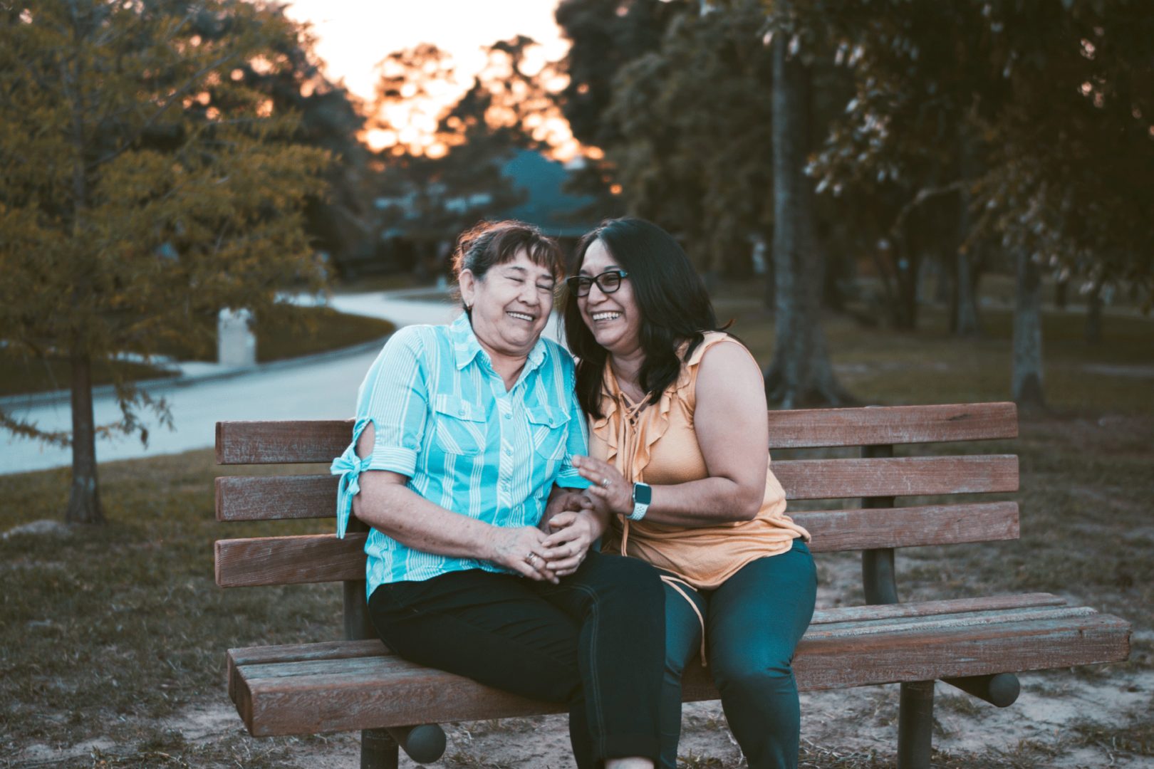 two elderly women sitting on a bench hug each other and smile