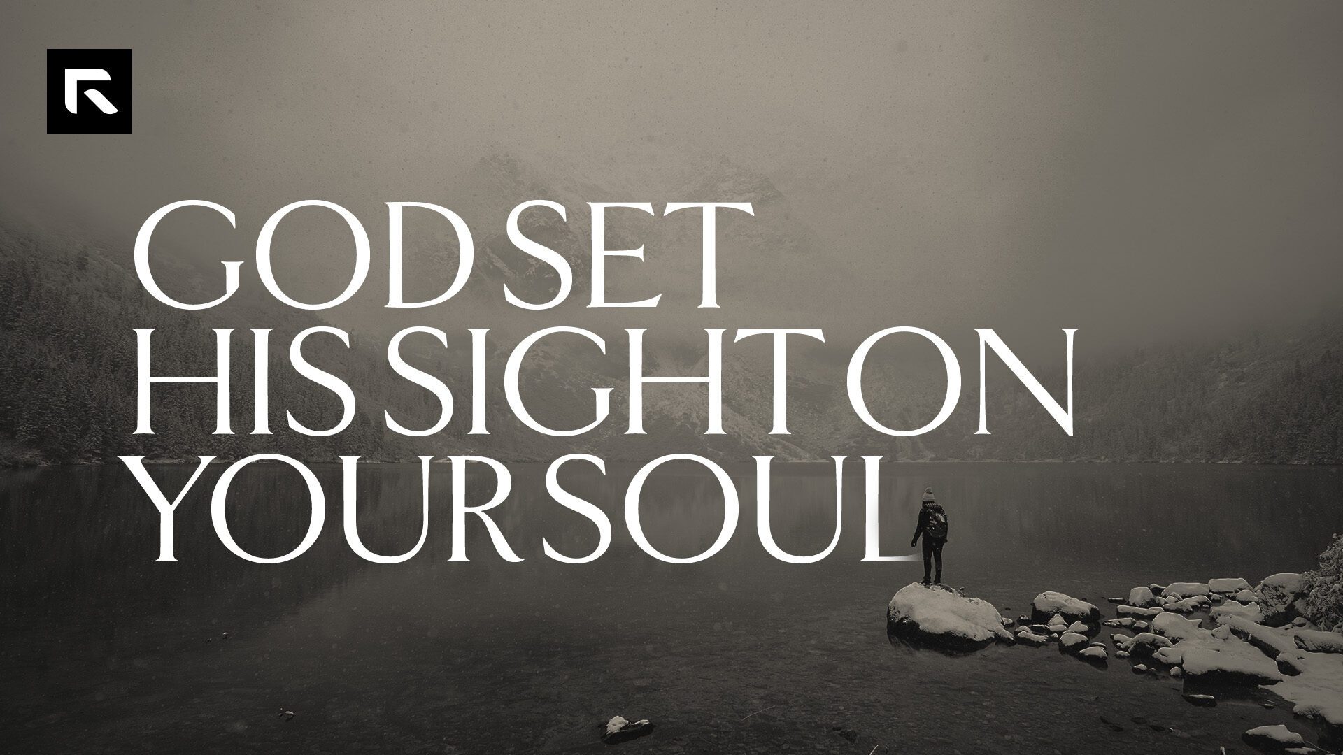 A balck and white picture of someone standing on a rock looking out to a lake and mountains covered in fog with the text "God Set His Sight On Your Soul"