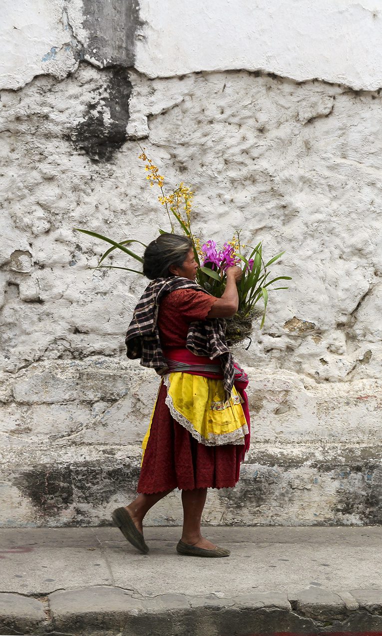 A woman carries a basket of flowers next to the cobblestone streets and crumbling walls of Antigua, Guatemala.