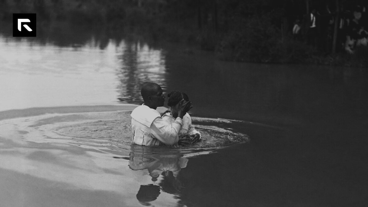 Black and white photo of a man wearing robes baptizing a women in a river