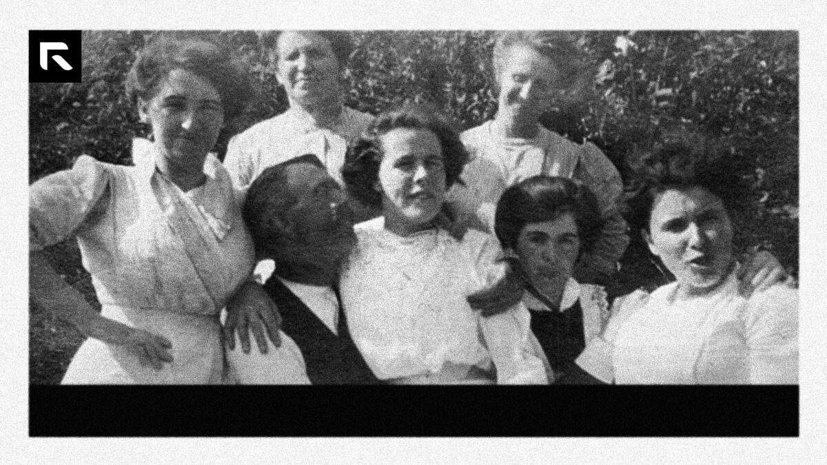 grainy black and white photo of a man sitting with six women gathered around him