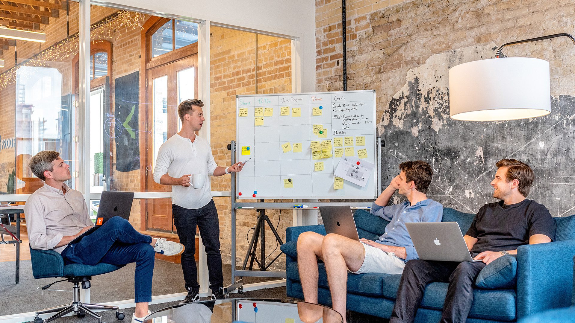 three men sitting using laptops while watching a fourth man explaining information on a whiteboard