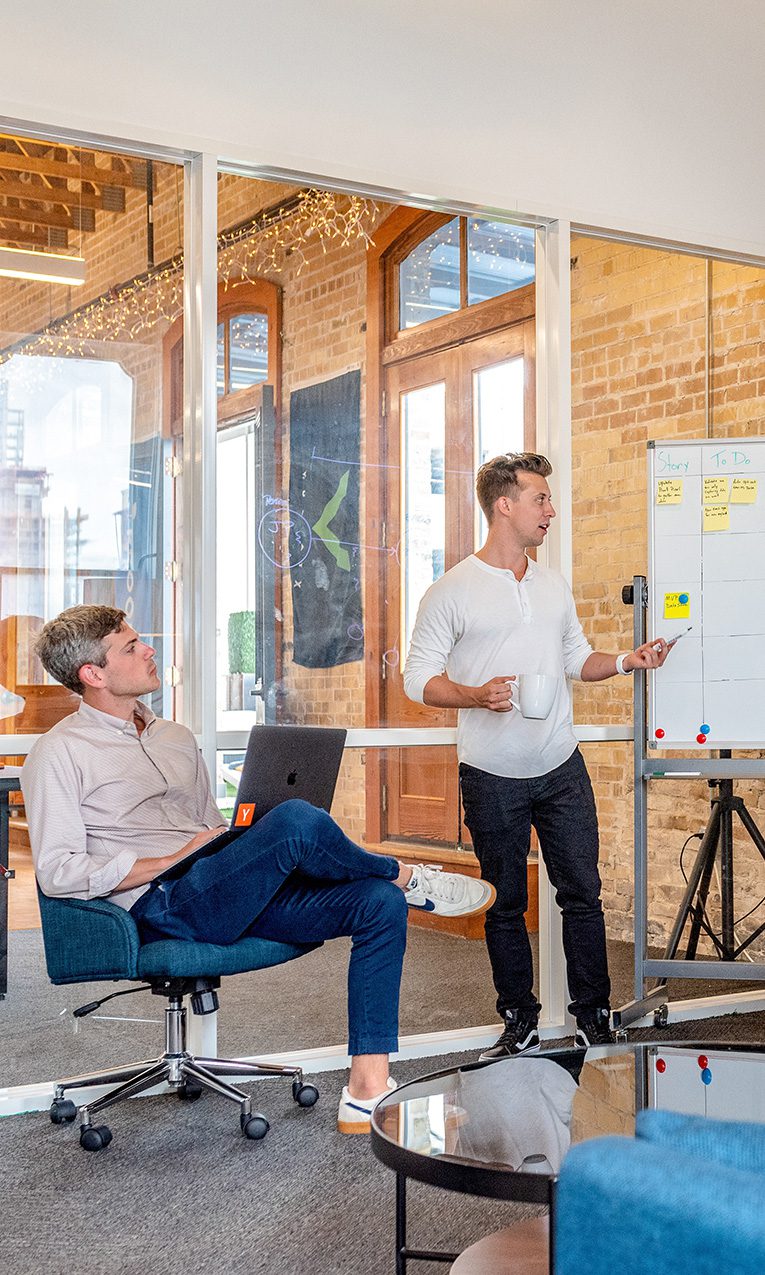three men sitting using laptops while watching a fourth man explaining information on a whiteboard