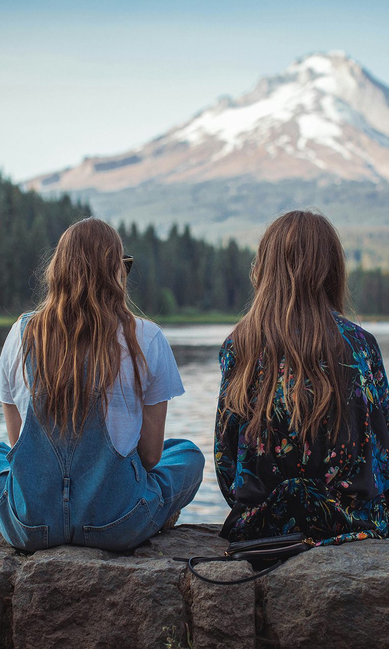 two women sitting on rock wall near body of water